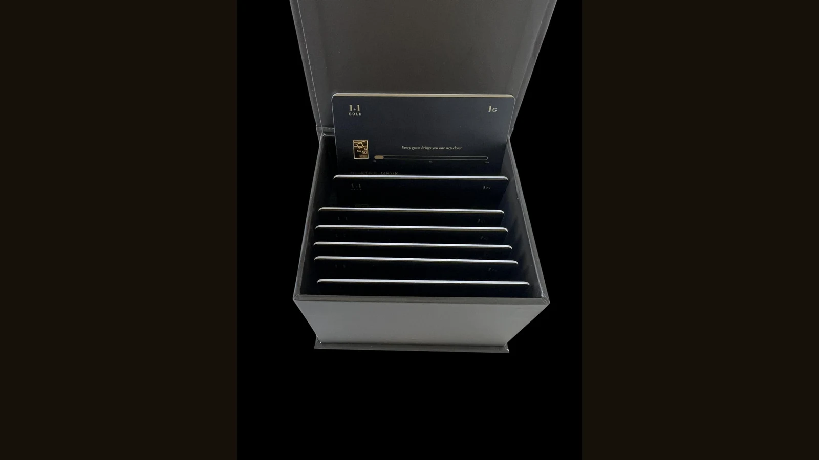 Five matte black 1Plus1Gold packaging cards fanned in a spread against a white background. The front card shows a 1g 999.9 fine gold bar seated in its window, the tagline 'Every gram brings you one step closer', a gold progress bar running from 0g to 100g, a laser-engraved serial number 1G-U6P8-Z25U, an NFC chip symbol, and the 1+1 Gold logo in gold lettering. Gold edges line each card.